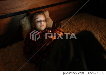 Top view of pretty child girl enjoying smartphone lying in bed, face illuminated by phone screen light. Preteen enjoying mobile apps, games, social media, online communication late at night. Top view of pretty child girl enjoying smartphone lying in bed, face illuminated by phone screen light. Preteen enjoying mobile apps, games, social media, online communication late at night. 122677118