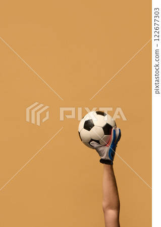 Hand wearing blue goalkeeper glove raising soccer ball with strong grip, focusing on goalkeeping skills and precision in football against beige background. 122677303