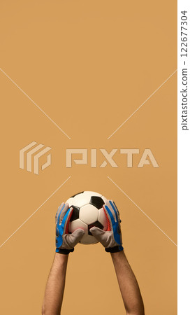 Both hands in blue goalkeeper gloves gripping soccer ball, highlighting teamwork and protection in competitive sports against beige background. 122677304