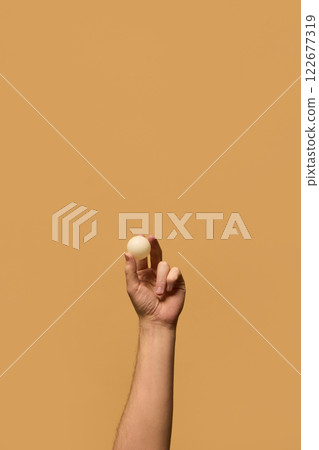 Hand holding small white ping pong ball between fingers, highlighting focus and control in precision-based table tennis matches against beige background. 122677319