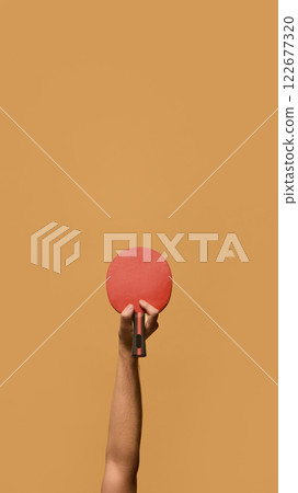 Hand lifting red table tennis paddle with strong grip, signifying confidence and skill in rapid ball exchanges against solid beige background. 122677320