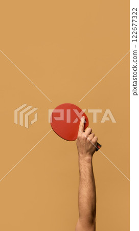 Hand raising red table tennis paddle with index finger extended, emphasizing technique and precision in competitive matches against beige background. Hand raising red table tennis paddle with index finger extended, emphasizing technique and precision in competitive matches against beige background. 122677322