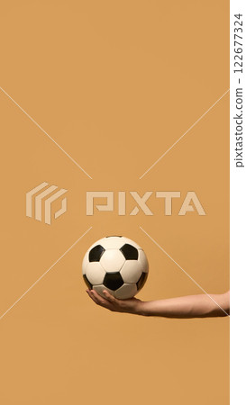Vertical image of hand holding soccer ball ready to throw it and start match against beige studio background. Energy essential for competitive play. 122677324