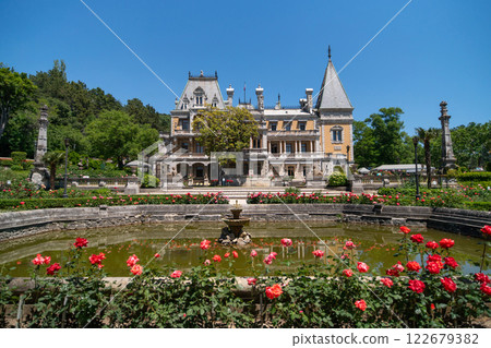 A large house with a fountain in front of it. The fountain is surrounded by red flowers. The house is surrounded by a garden with many trees and bushes. The sky is clear and blue. 122679382