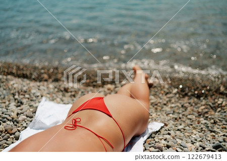 A woman sunbathes on the beach, lying on her stomach in a red swimsuit against the sea backdrop. A woman sunbathes on the beach, lying on her stomach in a red swimsuit against the sea backdrop. 122679413