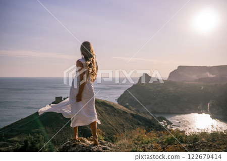 A woman in a white dress stands on a hill overlooking the ocean. The scene is serene and peaceful, with the woman's dress billowing in the wind. The combination of the ocean. A woman in a white dress stands on a hill overlooking the ocean. The scene is serene and peaceful, with the woman's dress billowing in the wind. The combination of the ocean. 122679414