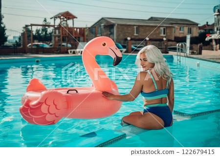 A woman is sitting on a pink inflatable flamingo in a pool. Concept of fun and relaxation, as the woman is enjoying her time in the water. A woman is sitting on a pink inflatable flamingo in a pool. Concept of fun and relaxation, as the woman is enjoying her time in the water. 122679415