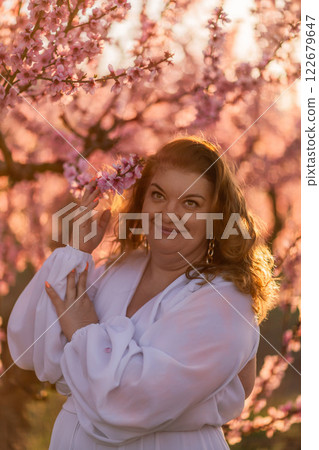 Woman blooming peach orchard. Against the backdrop of a picturesque peach orchard, a woman in a long white dress enjoys a peaceful walk in the park, surrounded by the beauty of nature. 122679647