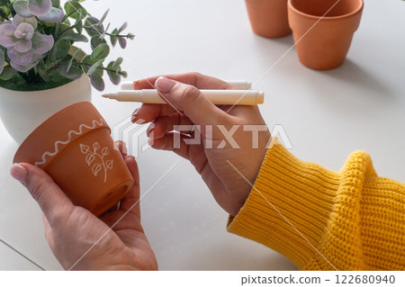 Caucasian woman decorating a terracotta pot with a white marker in a bright, cozy setting 122680940
