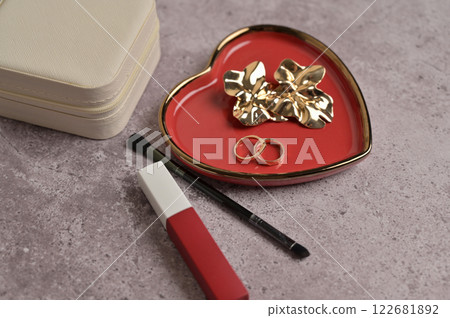 Women's trivia, cosmetic, couple of golden rings and earrings on red heart shape ceramic plate with a gold rim. Gray concrete background. Valentine's Day, marriage, wedding preparation 122681892
