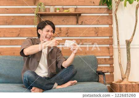 一個年輕的亞洲男子在家放鬆時用智慧型手機觀看體育比賽(握拳,快樂) 一個年輕的亞洲男子在家放鬆時用智慧型手機觀看體育比賽(握拳,快樂) 122685260