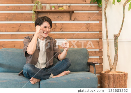 A young Asian man watching sports on his smartphone while relaxing at home (fist pump, happy) A young Asian man watching sports on his smartphone while relaxing at home (fist pump, happy) 122685262