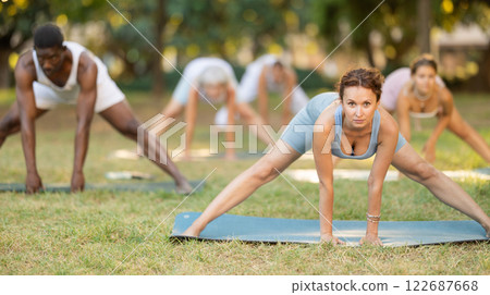 Multinational yoga enthusiasts with female coach perform Prasarita Padottanasana in park Multinational yoga enthusiasts with female coach perform Prasarita Padottanasana in park 122687668