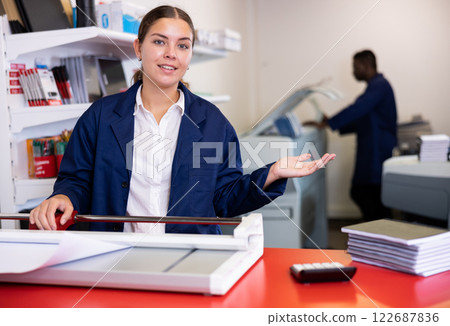 Portrait of cheerful young female typographer in uniform using paper cutter during work in printer shop 122687836