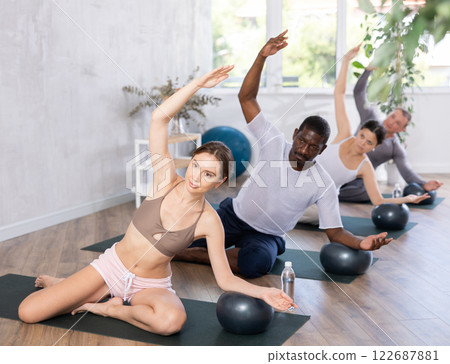 Woman in activewear sitting on yoga mat during group workout with fitness ball at gym. Healthy lifestyle concept Woman in activewear sitting on yoga mat during group workout with fitness ball at gym. Healthy lifestyle concept 122687881