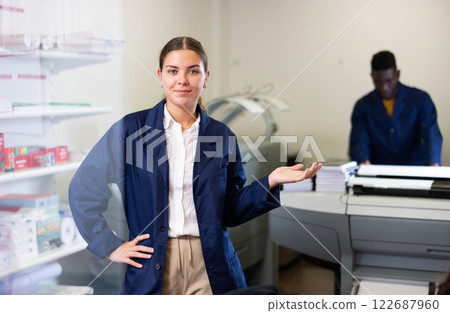Cheerful young woman in blue robe uniform sticking out her left hand to show the workflow in the printer house 122687960