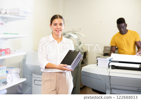 Portrait of happy young female specialist holding printed samples of notebooks in the print shop Portrait of happy young female specialist holding printed samples of notebooks in the print shop 122688013