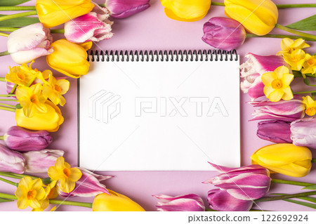 Open paper notebook with empty page with purple and yellow tulips and yellow daffodils on purple background. Top view, flat lay, work space on purple background. Open paper notebook with empty page with purple and yellow tulips and yellow daffodils on purple background. Top view, flat lay, work space on purple background. 122692924