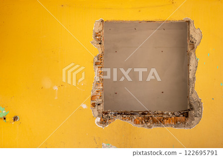 Close-up of a square hole in a yellow wall, revealing a patch of grey concrete underneath. Shows damage, repair, or renovation in progress.  122695791