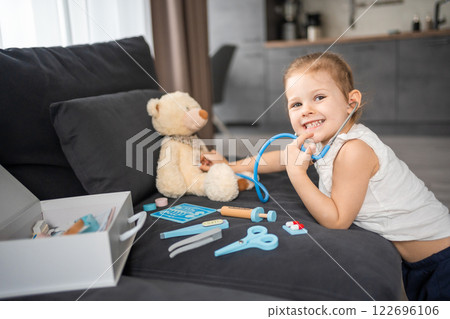 Little girl playing doctor with toys and teddy bear on the sofa in living room at home 122696106