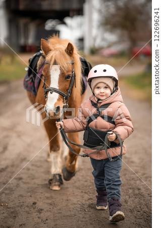 Little girl in protective jacket and helmet with her brown pony before riding Lesson 122696241