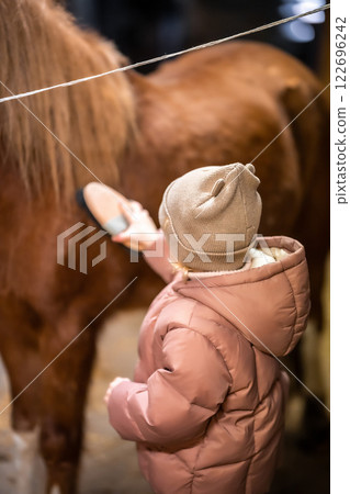 Horse care inside the stable before the ride. Little cute girl and pony. 122696242