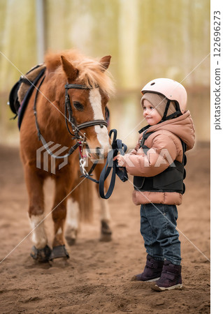 Portrait of little girl in protective jacket and helmet with her brown pony before riding Lesson Portrait of little girl in protective jacket and helmet with her brown pony before riding Lesson 122696273