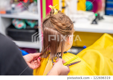 Close up view of hairdresser hands making a hair style to cute little girl in real child salon with colorful interior and toys Close up view of hairdresser hands making a hair style to cute little girl in real child salon with colorful interior and toys 122696319