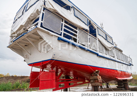 Stern of large boat on wooden supports for repair on dry dock, with covered upper deck and metal railings, low angle view against cloudy sky Stern of large boat on wooden supports for repair on dry dock, with covered upper deck and metal railings, low angle view against cloudy sky 122696331