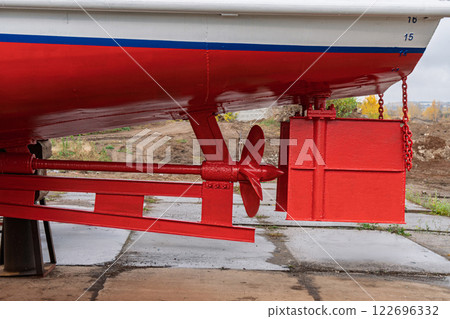 Dry-docked boat with a red and white hull, showing its rudder system and propeller, supported by wooden and metal blocks, in shipyard close view Dry-docked boat with a red and white hull, showing its rudder system and propeller, supported by wooden and metal blocks, in shipyard close view 122696332