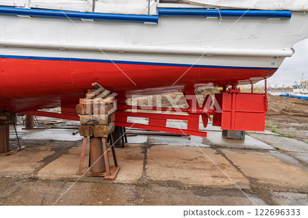 Dry-docked boat with a red and white hull, showing its rudder system and propeller, supported by wooden and metal blocks, in shipyard close view Dry-docked boat with a red and white hull, showing its rudder system and propeller, supported by wooden and metal blocks, in shipyard close view 122696333