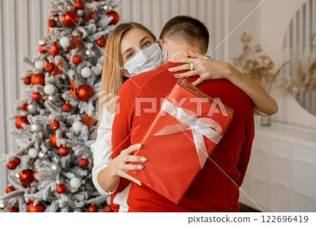 Close up of happy couple in medical masks with christmas gift hugging at home near Christmas tree Close up of happy couple in medical masks with christmas gift hugging at home near Christmas tree 122696419