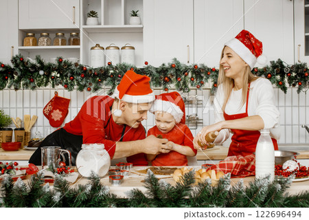 Happy parents in santa hats and little son are in the kitchen preparing dough for baking Christmas cookies. Having fun, playing, a happy family waiting for the holidays. 122696494