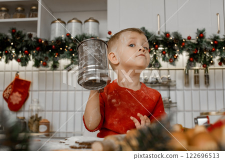 Happy fair-haired child in red sweater stands at the kitchen table with iron sieve in his hand at home looking proudly into camera. 122696513