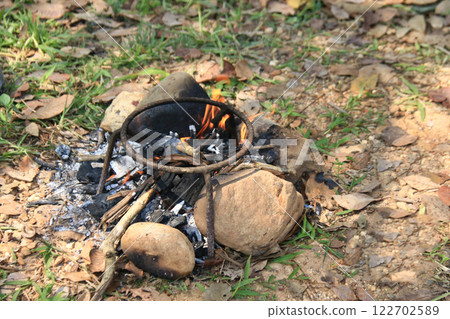 A classic three legged iron stand is placed over an open fire, designed to hold cooking pots. This method reflects traditional outdoor cooking, commonly used in camping and rural kitchens. A classic three legged iron stand is placed over an open fire, designed to hold cooking pots. This method reflects traditional outdoor cooking, commonly used in camping and rural kitchens. 122702589