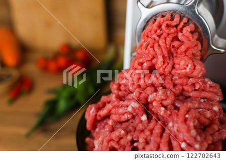Concept of healthy and proper nutrition, cooking.Close-up of red ground beef with fat streaks coming out of a metal meat grinder on a wooden background, vegetables and herbs are visible out of focus 122702643