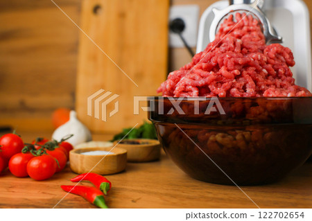Side view on a wooden background: dark dishes with red minced meat inside, which comes out of a metal meat grinder, spices, herbs and vegetables are nearby. The concept of healthy eating, cooking 122702654
