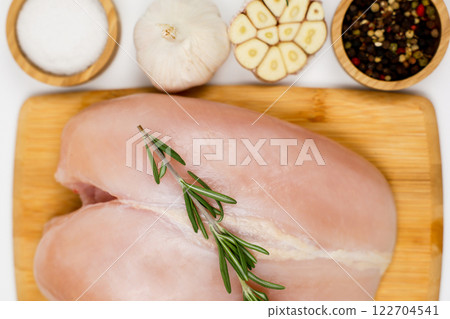 A close-up view from above of chicken breast fillet with a sprig of rosemary lying on a wooden board, nearby are wooden bowls with pepper, seasoning and salt and garlic on a white background. The A close-up view from above of chicken breast fillet with a sprig of rosemary lying on a wooden board, nearby are wooden bowls with pepper, seasoning and salt and garlic on a white background. The 122704541