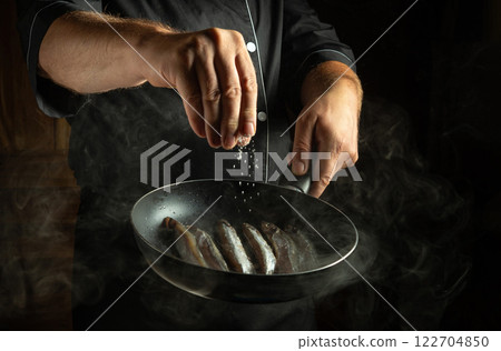 A skilled chef adds a sprinkle of salt over a sizzling pan filled with fresh fish. The dark and smoky ambiance enhances the art of culinary preparation, showcasing expertise and passion. 122704850