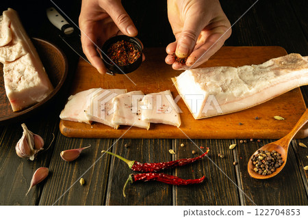Hands carefully sprinkle spices over slices of pork belly, surrounded by garlic and chili peppers on a wooden table. The warm light creates a cozy atmosphere perfect for culinary creativity 122704853
