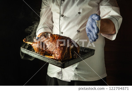 A skilled chef showcases a beautifully roasted duck on a black tray, with smoke rising from the dish. The warm lighting highlights the rich color of the cooked meat in a welcoming kitchen 122704871