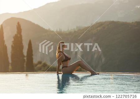 Slim woman in swimsuit walking by the edge of luxury infinity pool with jungle view Slim woman in swimsuit walking by the edge of luxury infinity pool with jungle view 122705716