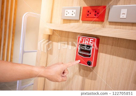 Hand pointing at red fire alarm switch on wooden wall in the office interior with electric sockets Hand pointing at red fire alarm switch on wooden wall in the office interior with electric sockets 122705851