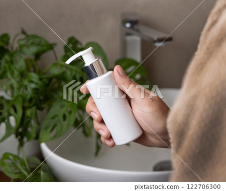 Woman in brown bathrobe with a white cream bottle in hand in bathroom closeup, cosmetic mockup 122706300