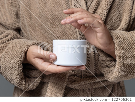 Closeup of woman in brown bathrobe holding white cream jar in hands, cosmetic mockup Closeup of woman in brown bathrobe holding white cream jar in hands, cosmetic mockup 122706301