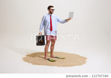 Man standing on sand wearing flippers, holding briefcase and laptop, wearing sunglasses, formal shirt, tie, swim trunks against white studio background. Man standing on sand wearing flippers, holding briefcase and laptop, wearing sunglasses, formal shirt, tie, swim trunks against white studio background. 122707160