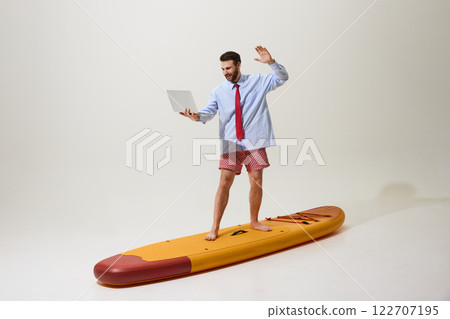Businessman in formal shirt, tie, and shorts standing on paddleboard with laptop, waving confidently against white studio background. 122707195