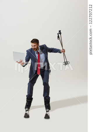 Businessman in suit and red tie balancing on skis, holding laptop in one hand and ski poles in other, smiling against white studio background. Businessman in suit and red tie balancing on skis, holding laptop in one hand and ski poles in other, smiling against white studio background. 122707212