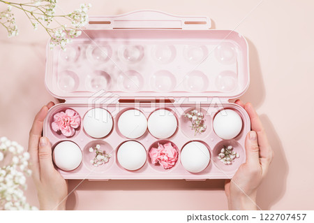 Female hands holding pink egg carton filled with white eggs and delicate pink flowers on pastel pink background. Soft and creative design perfect for holiday Easter and spring-inspired themes Female hands holding pink egg carton filled with white eggs and delicate pink flowers on pastel pink background. Soft and creative design perfect for holiday Easter and spring-inspired themes 122707457