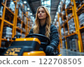 Female warehouse worker drives yellow forklift in an industrial storage facility with tall racks filled with goods 122708505